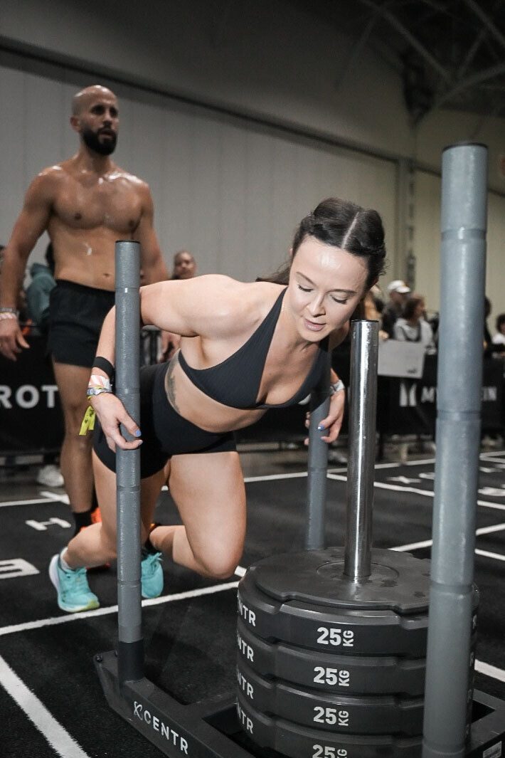 Alana driving a weighted sled forward, one knee down, during her HYROX race